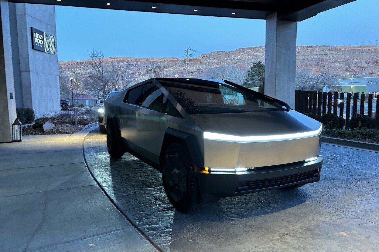 A wrecked silver Tesla Cybertruck with a crushed rear bed and damaged rear wheel, parked in an auction yard lot.